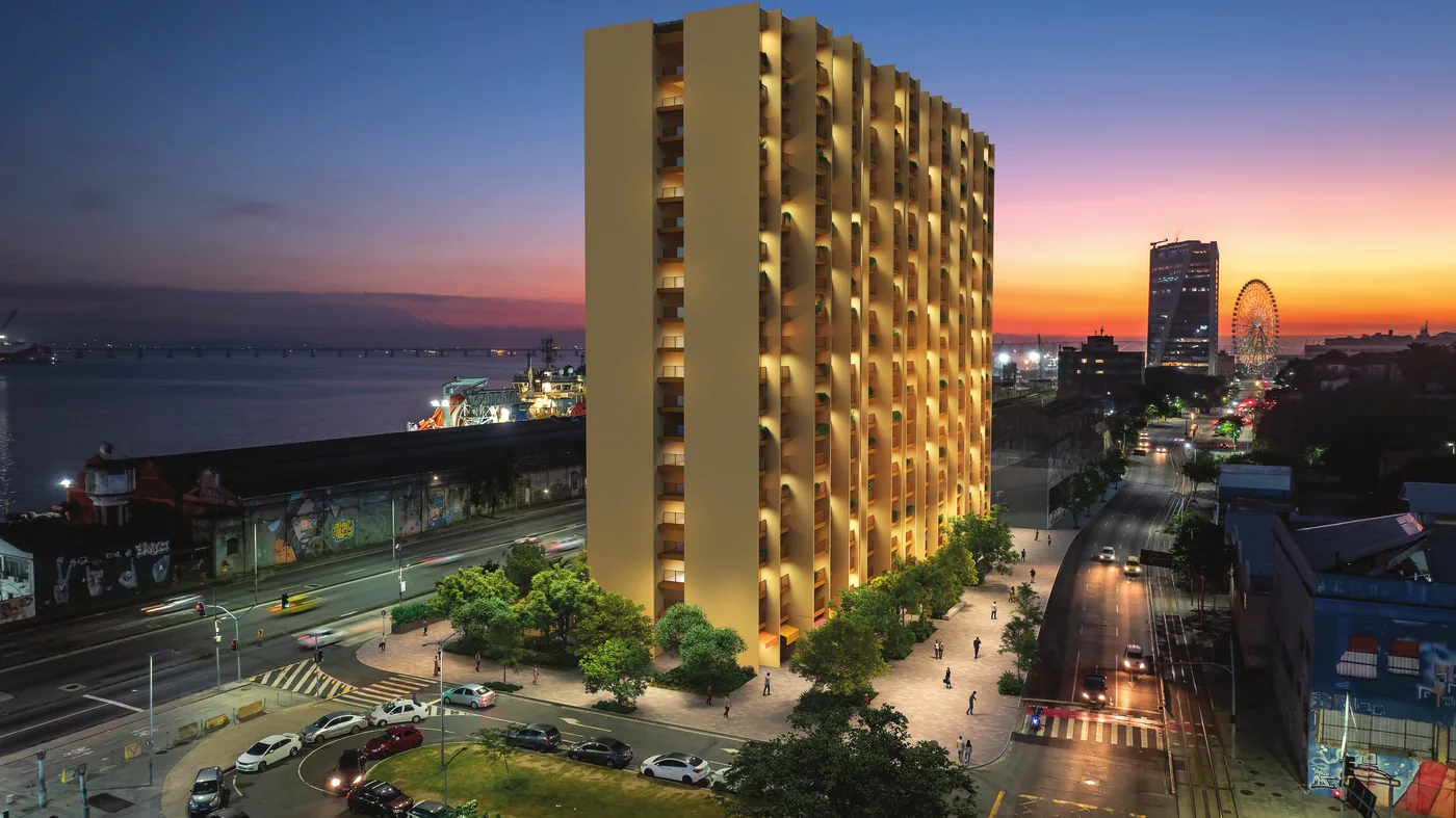 Fachada do Residencial Erê ao anoitecer, na Avenida Rodrigues Alves, em Santo Cristo, com a Baía de Guanabara e a roda-gigante Yup Star ao fundo